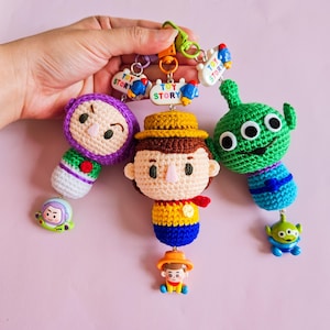 May include: Three crocheted Toy Story characters: a purple and green Buzz Lightyear, a brown and yellow Woody, and a green alien with three eyes. Each character has a smaller version of themselves attached to the bottom. The characters are attached to a keychain with a "Toy Story" charm.