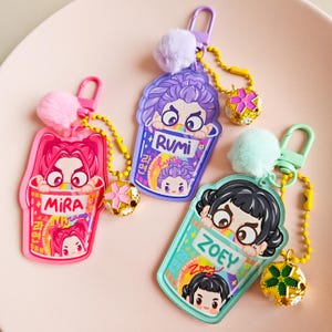 May include: Three vibrant acrylic keyrings with cartoon character designs. Each keyring features a different character name: Mira, Rumi, and Zoey. They have a pom-pom and a decorative charm attached to a chain.