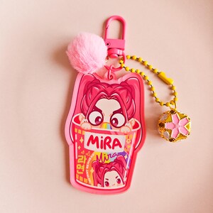 May include: A pink keychain featuring a cartoon girl in a ramen bowl design. It includes a pink pom-pom, a gold beaded chain, and a charm with a pink flower. The word "Mira" is printed on the bowl, and the background is pink.