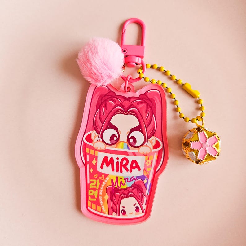 May include: A pink keychain featuring a cartoon girl in a ramen bowl design. It includes a pink pom-pom, a gold beaded chain, and a charm with a pink flower. The word "Mira" is printed on the bowl, and the background is pink.