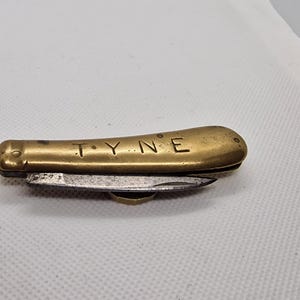 May include: A small, brass pocket knife with a single blade. The knife is closed and has the word "TYNE" engraved on the handle.