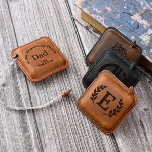 Könnte beinhalten: Vier Ledermaßbänder mit personalisierten Gravuren. Die Bänder sind braun und haben verschiedene Designs, darunter ein Monogramm, ein Lorbeerkranz mit einem Buchstaben und eine Botschaft, die lautet "No One Measures Up To You Dad Love Wyatt & Grace".