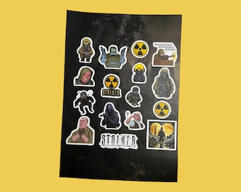 Puede incluir: Una hoja de pegatinas con personajes y símbolos del videojuego Stalker. Las pegatinas están sobre un fondo negro e incluyen una variedad de personajes, como un soldado, un científico y un mutante. Las pegatinas también presentan el logotipo del juego y un símbolo de radiación.