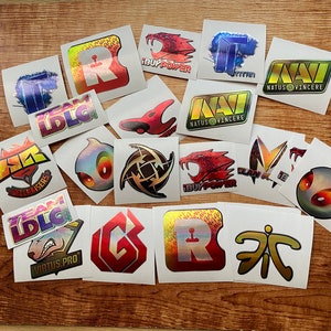 May include: A collection of 15 colorful holographic stickers featuring various esports team logos, including Team Liquid, Virtus.pro, Natus Vincere, and iBUYPOWER. The stickers are on a wooden surface.