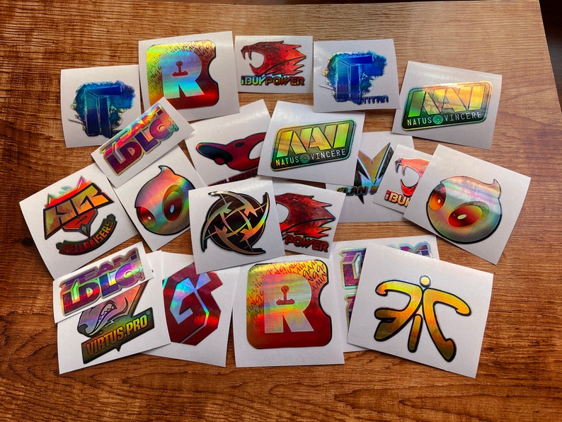 May include: A collection of 16 holographic stickers featuring various esports team logos, including Virtus.pro, Team Liquid, Natus Vincere, and iBUYPOWER. The stickers are on a wooden surface.