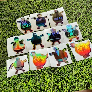 May include: A set of 10 iridescent holographic stickers featuring various characters and designs. The stickers are on white cardstock backgrounds. The characters include a chicken, a person wearing a hat, a person wearing a vest, and a person wearing a suit.