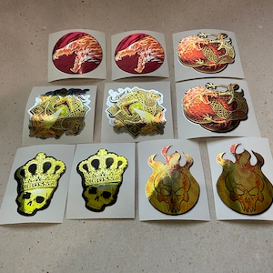 May include: Ten assorted holographic stickers featuring a variety of designs, including dragons, skulls, and flames. The stickers are all gold and red with a holographic finish.