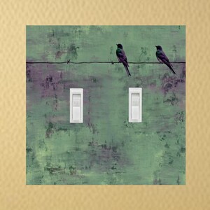 May include: A square wall art print featuring two black birds perched on a wire against a textured green and purple background. Two white light switch covers are below the wire. The artwork is mounted on a gold-colored wall.