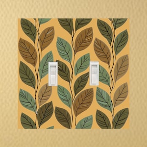 May include: A decorative light switch cover featuring a repeating pattern of stylized leaves in shades of brown, green, and blue-green against a mustard yellow background. Two white light switches are visible. The design adds a touch of nature-inspired decor.