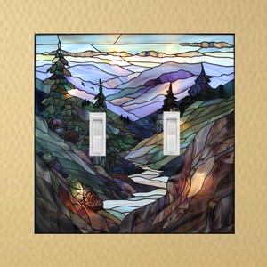 May include: A stained glass light switch cover featuring a mountain landscape with a river. The artwork uses shades of blue, green, and brown. Two white light switches are centered on the cover. The cover is square and mounted on a gold wall.
