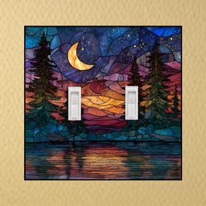 May include: A decorative light switch cover featuring a stained-glass design of a night scene. The artwork depicts a crescent moon, starry sky, pine trees, and a lake with reflections. The cover has two white light switches.
