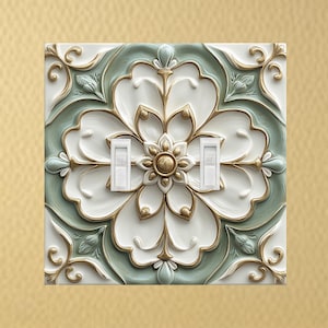 May include: A decorative light switch cover featuring a floral design. The cover is square, with a white, green, and gold color scheme. The floral pattern is raised, creating a 3D effect. Two white light switches are visible.
