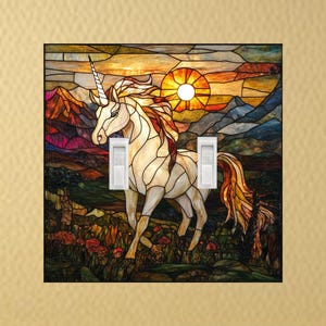 May include: A stained glass light switch cover featuring a white unicorn with a flowing mane and tail. The background depicts a sunset over mountains and a field of flowers. The cover has two white light switch toggles.