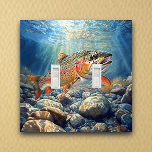 May include: A light switch cover featuring a colorful trout swimming through a rocky stream bed. The trout is facing the viewer with its mouth open. The water is clear and blue, and the rocks are brown and gray. The light switch cover is a standard size and is suitable for most light switches.