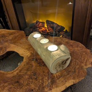 May include: A wooden candle holder with three tealight candles burning. The holder is made from a natural log and has a rustic look. It is sitting on a wooden table with a natural wood grain.