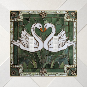 May include: A square double light switch cover plate featuring a stained glass style design of two white swans facing each other against a dark green background. The plate has two white toggle switches and a decorative metal border frame.