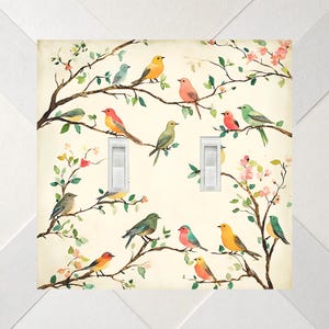 May include: A double light switch wall plate featuring a cream-colored background with a pattern of various colorful birds perched on brown tree branches with small green leaves and pink blossoms. Two white toggle switches are centered on the plate.