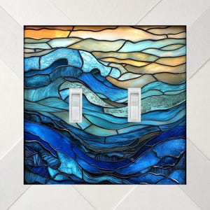 May include: A double light switch cover plate featuring a stained glass style design of ocean waves in shades of deep blue, turquoise, and light blue, with a golden sunset sky at the top. Two white toggle switches are centered on the plate.