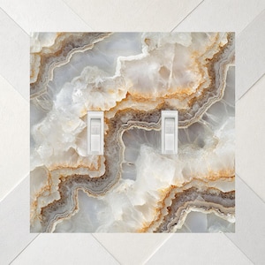 May include: A double light switch wall plate featuring a printed pattern of white, cream, and amber-toned agate stone with wavy, layered mineral bands. The rectangular plate has two vertical white toggle switches centered on the surface.