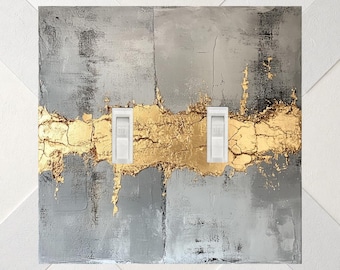 May include: A double light switch wall plate featuring a textured gray background with a horizontal band of metallic gold leaf across the center. Two white toggle switches are vertically aligned within the gold section of the plate.