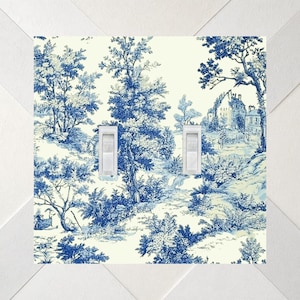 May include: A double light switch cover plate featuring a blue and white toile pattern. The design depicts a landscape with trees, foliage, and buildings. Two white toggle switches are centered vertically on the rectangular plate.