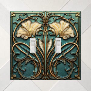 May include: A double light switch cover plate featuring a teal background with embossed gold filigree and cream-colored floral motifs. The rectangular plate has two vertical white toggle switches centered within the ornate, symmetrical design.