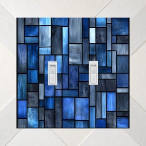 May include: A double light switch wall plate featuring a rectangular grid pattern of various shades of blue and grey, resembling stained glass. The design consists of vertical and horizontal blocks with two white toggle switches in the center.