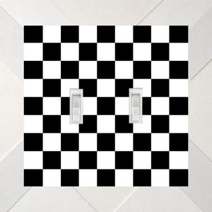 May include: A square double light switch wall plate featuring a high-contrast black and white checkerboard pattern. The plate includes two vertical white toggle switches positioned side-by-side in the center of the checkered surface.
