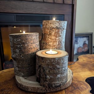 May include: Three wooden candle holders with lit tea lights on a wooden surface. The candle holders are made from birch logs and have a natural bark finish.