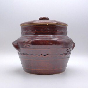 May include: A brown ceramic crock with a lid. The crock has a round body with two handles and is decorated with wave and star patterns. The lid has a small knob. The crock is likely used for food storage.