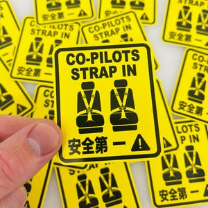 以下が含まれることがあります： 黒い文字とグラフィックが入った黄色の正方形のステッカー。「CO-PILOTS STRAP IN」の文字が、シートベルト付きの2つの座席のグラフィックの上に書かれています。下には別の言語のテキストがあります。ステッカーは白い表面に散らばっています。