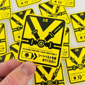 Może przedstawiać: Żółto-czarna naklejka z grafiką pasów bezpieczeństwa i japońskim tekstem. Naklejka głosi "PLEASE FASTEN YOUR SEATBELT" i "シートベルトを締めてください". Naklejka ma symbol ostrzegawczy i słowo "注意".