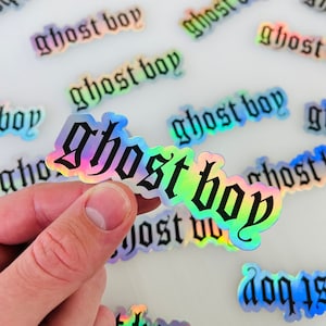 Puede incluir: Una colección de pegatinas holográficas iridiscentes con el texto "ghost boy" en una fuente gótica. Las pegatinas están esparcidas sobre una superficie blanca.