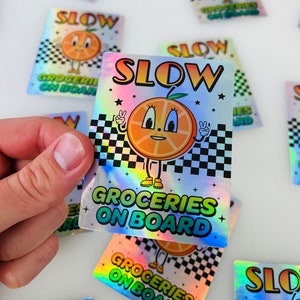 Puede incluir: Una pegatina holográfica con una naranja de dibujos animados haciendo un signo de paz y el texto "SLOW GROCERIES ON BOARD". La pegatina tiene un fondo a cuadros y estrellas.