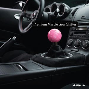 Può includere: Un pomello del cambio in marmo rosa all'interno di un'auto. Il pomello è una sfera con un motivo a vortice di bianco e rosa. Il testo "Premium Marble Gear Shifter" è sopra il pomello.