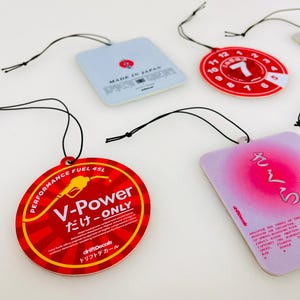 May include: Four car air fresheners with red and white designs. One air freshener is round and says "V-Power だけ-ONLY" with a petrol pump graphic. Another air freshener is round and has a red number 7 with a white background. The third air freshener is square and says "MADE IN JAPAN". The fourth air freshener is square and has a pink background with Japanese text that says "さくら".