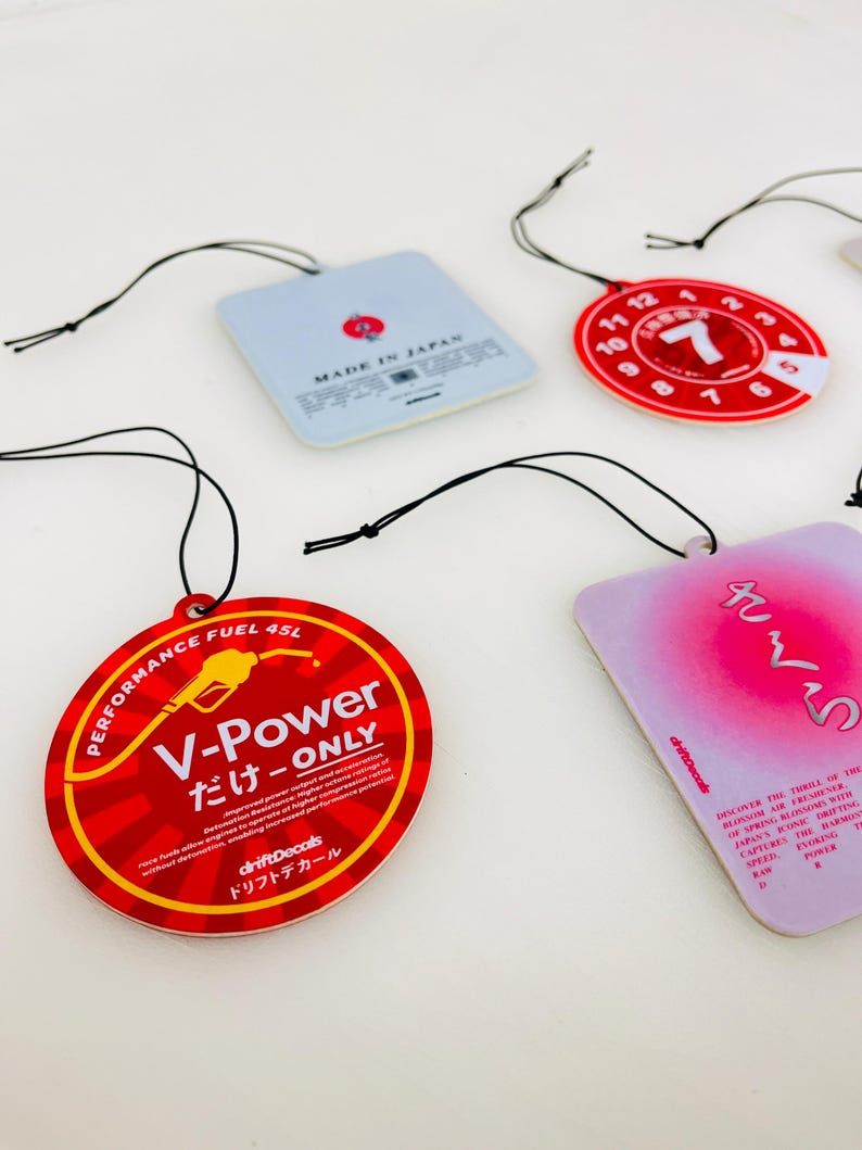 May include: Four car air fresheners with red and white designs. One air freshener is round and says "V-Power だけ-ONLY" with a petrol pump graphic. Another air freshener is round and has a red number 7 with a white background. The third air freshener is square and says "MADE IN JAPAN". The fourth air freshener is square and has a pink background with Japanese text that says "さくら".