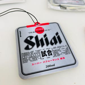 May include: A silver car air freshener with a black and red design. The text "Shiai" is printed in black and red, and the text "Super Performance" is printed in red. The text "試合" is printed in black. The text "スーパー パフォーマンス 試合" is printed in black. The text "200ml" is printed in black.