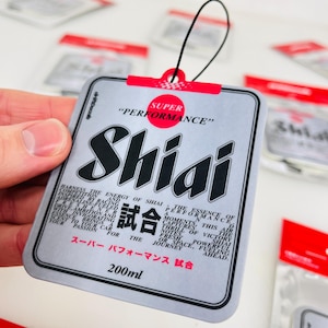 Op de afbeelding: Een zilveren autoluchtverfrisser met een zwart-wit ontwerp. De luchtverfrisser heeft de tekst "Shiai" en "Super Performance" in rood en zwart. De tekst "試合" staat ook op de luchtverfrisser. De luchtverfrisser is 200 ml.