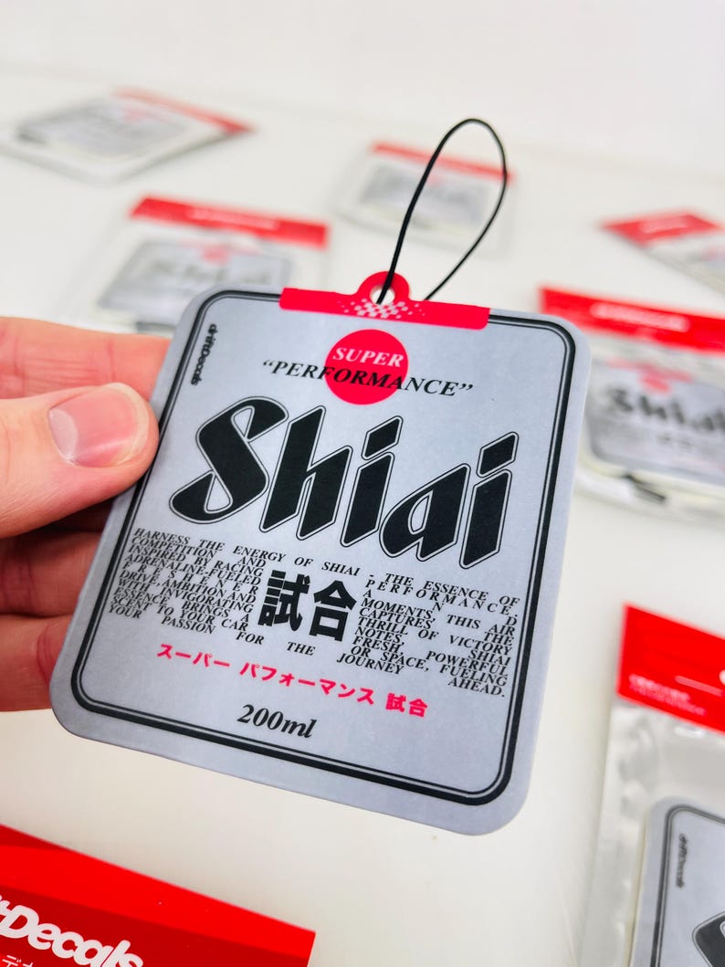 May include: A silver car air freshener with a black and white design. The air freshener has the text "Shiai" and "Super Performance" in red and black. The text "試合" is also on the air freshener. The air freshener is 200 ml.