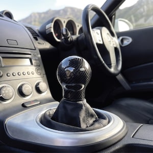 May include: A spherical carbon fibre gear shift knob with a glossy finish, mounted on a black leather shift boot within a vehicle interior. The gear shifter is positioned on a silver-toned centre console near the dashboard and steering wheel.