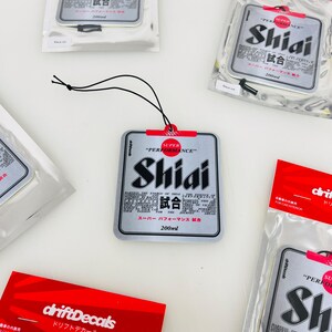 May include: A silver car air freshener with a red and white label that says "Super Performance Shiai 試合" and "200ml". The air freshener is hanging from a black string.