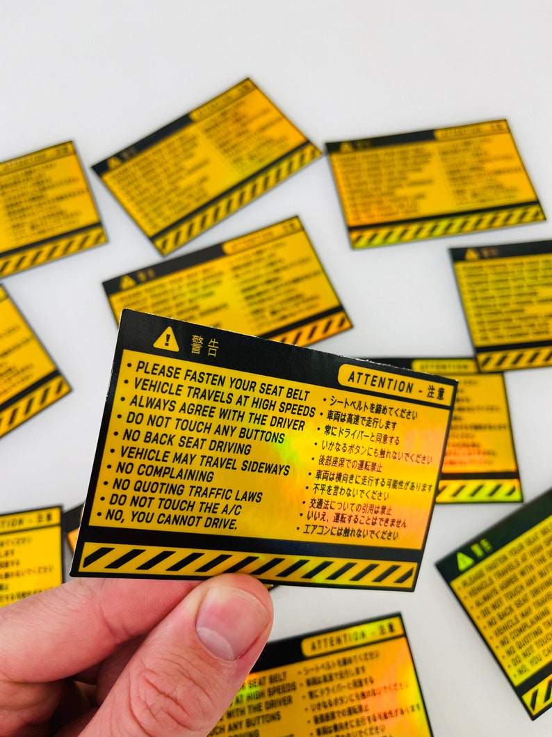 May include: A yellow and black warning sticker with a holographic finish. The sticker has a black exclamation point and the text "ATTENTION - 注意" in Japanese and English. The text reads: "Please fasten your seat belt. Vehicle travels at high speeds. Always agree with the driver. Do not touch any buttons. No back seat driving. Vehicle may travel sideways. No complaining. No quoting traffic laws. Do not touch the A/C. No, you cannot drive."