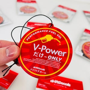 May include: A round car air freshener in red with a black string for hanging. It features a yellow petrol pump with a red nozzle and the text "PERFORMANCE FUEL 45L V-Power だけ-ONLY" in white and black. The text "driftDecals ドリフトデカール" is in white and black on a red background.