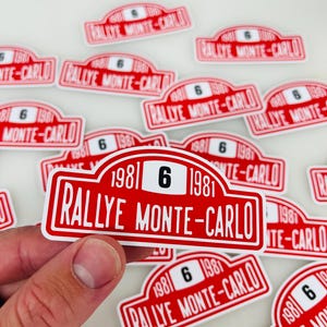 May include: Red and white stickers with the text "RALLYE MONTE-CARLO 1981 6". The stickers are rectangular with rounded corners and a white border. The number 6 is in black.