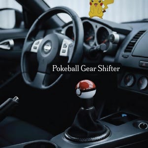 Op de afbeelding: Auto-interieur met een Pokeball versnellingspook, rood en wit van kleur. Een Pikachu luchtverfrisser hangt aan de spiegel. De tekst "Pokeball Gear Shifter" is zichtbaar.