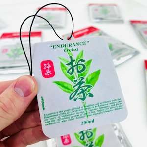 May include: White car air freshener with green tea leaves and Japanese characters. The text "ENDURANCE" and "Ocha" are printed on the front. The air freshener is 200 ml.