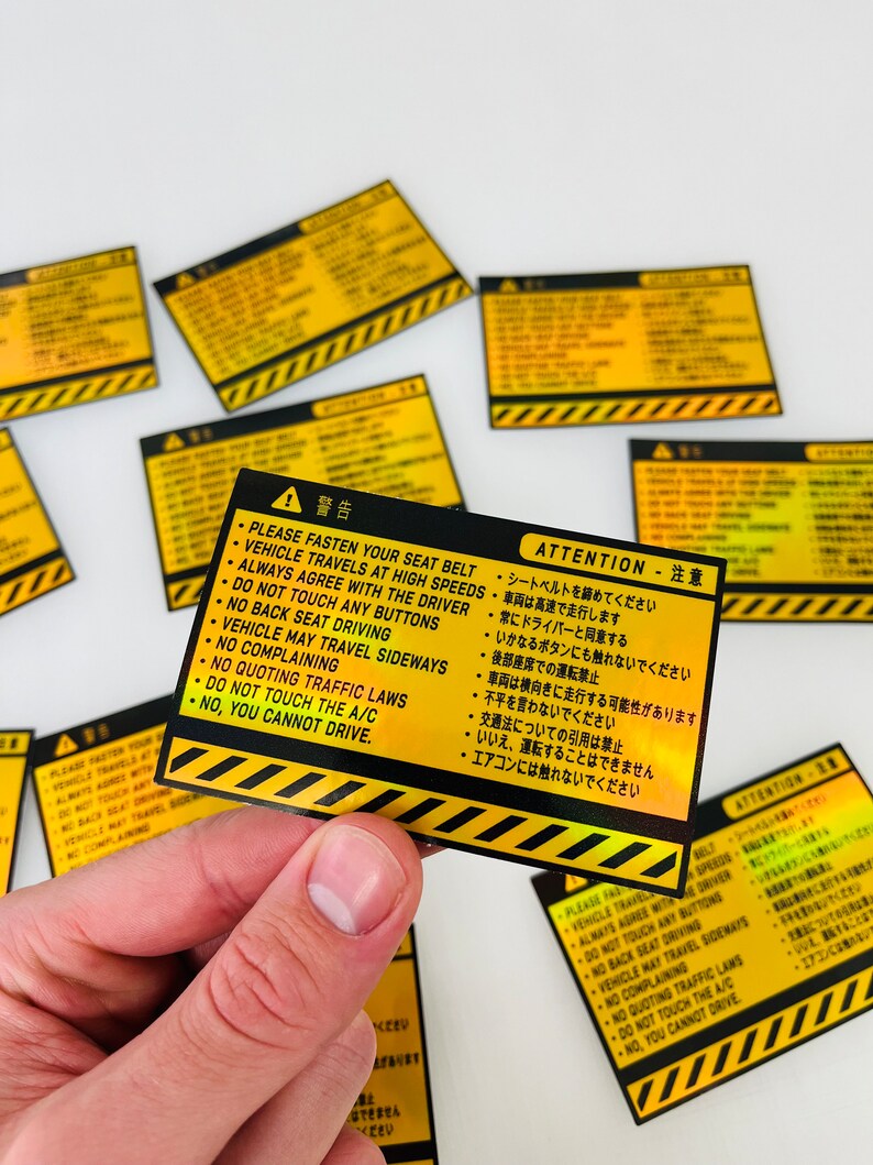 May include: A yellow and black warning sign with a black triangle and exclamation point. The sign has Japanese and English text that reads: "Attention - Please fasten your seat belt. Vehicle travels at high speeds. Always agree with the driver. Do not touch any buttons. No back seat driving. Vehicle may travel sideways. No complaining. No quoting traffic laws. Do not touch the A/C. No, you cannot drive."
