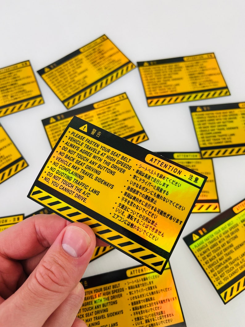 May include: A yellow and black warning sticker with a red exclamation point and the text "ATTENTION - 注意" in Japanese. The sticker has a list of rules for passengers in a vehicle, including "Please fasten your seat belt", "Vehicle travels at high speeds", "Always agree with the driver", "Do not touch any buttons", "No back seat driving", "Vehicle may travel sideways", "No complaining", "No quoting traffic laws", "Do not touch the A/C", and "No, you cannot drive."
