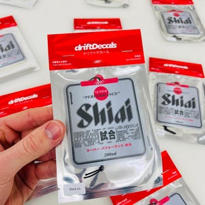 May include: A silver car air freshener with the text "Shiai" and "Super Performance" in black and red. The air freshener is packaged in a clear plastic bag with a black string.
